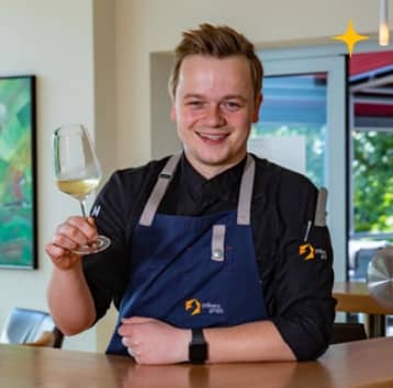 Felix Feldkamp - ein junger Mann mit Kochschürze hält einWeißweinglas hoch und lächelt in die Kamera