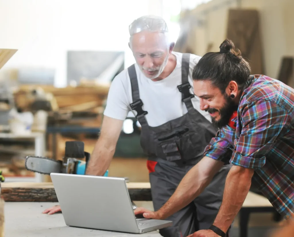 Mann in Handwerker-Latzhose und Mann in einem bunt-karierten Hemd stehe in der Werkstatt und schauen gemeinsam auf einen Laptop